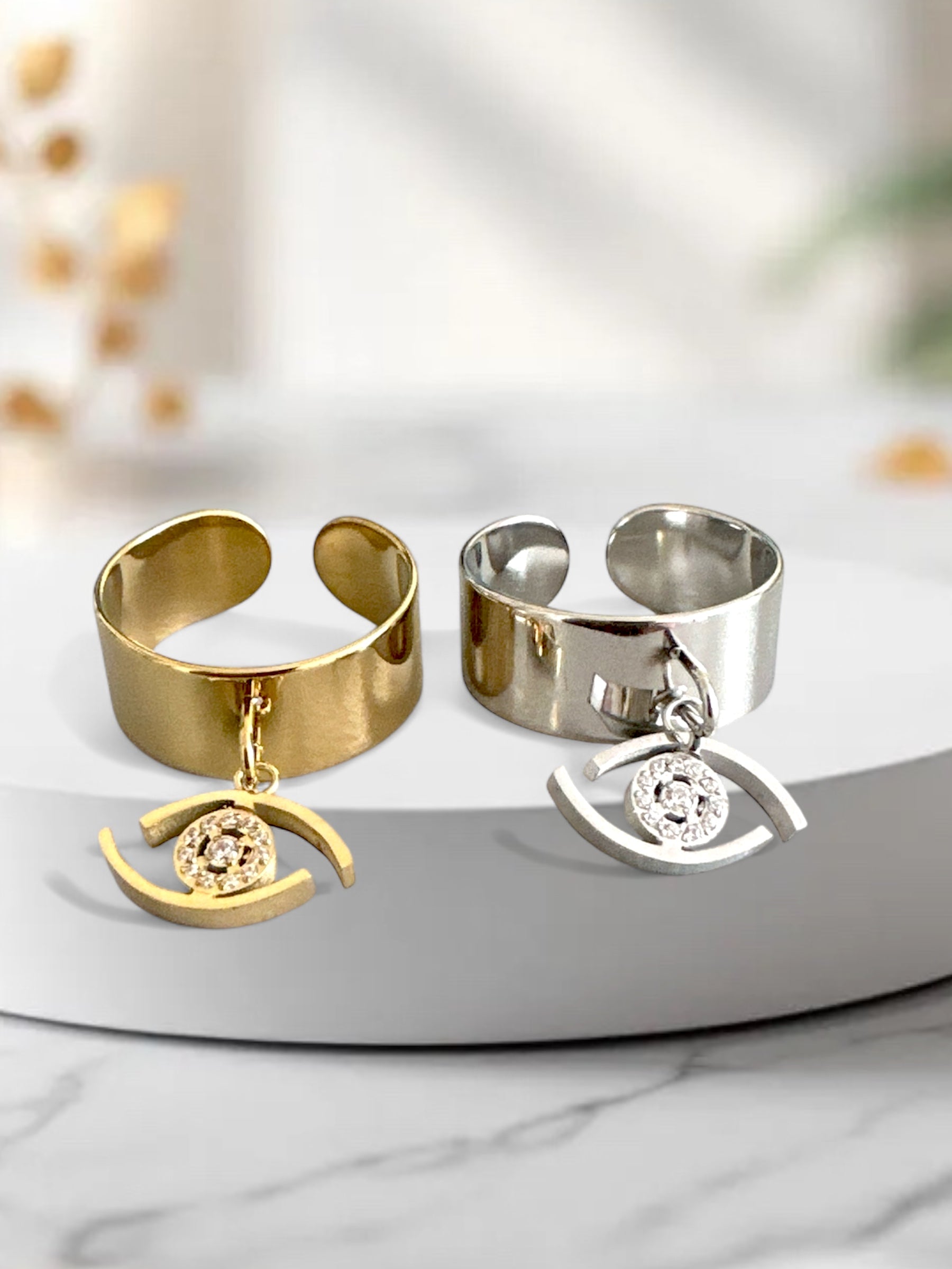 Ατσάλινα δαχτυλίδια σε χρυσό και ασημί με κρεμαστό charm σε σχήμα ματιού και λαμπερή πέτρα στο κέντρο.