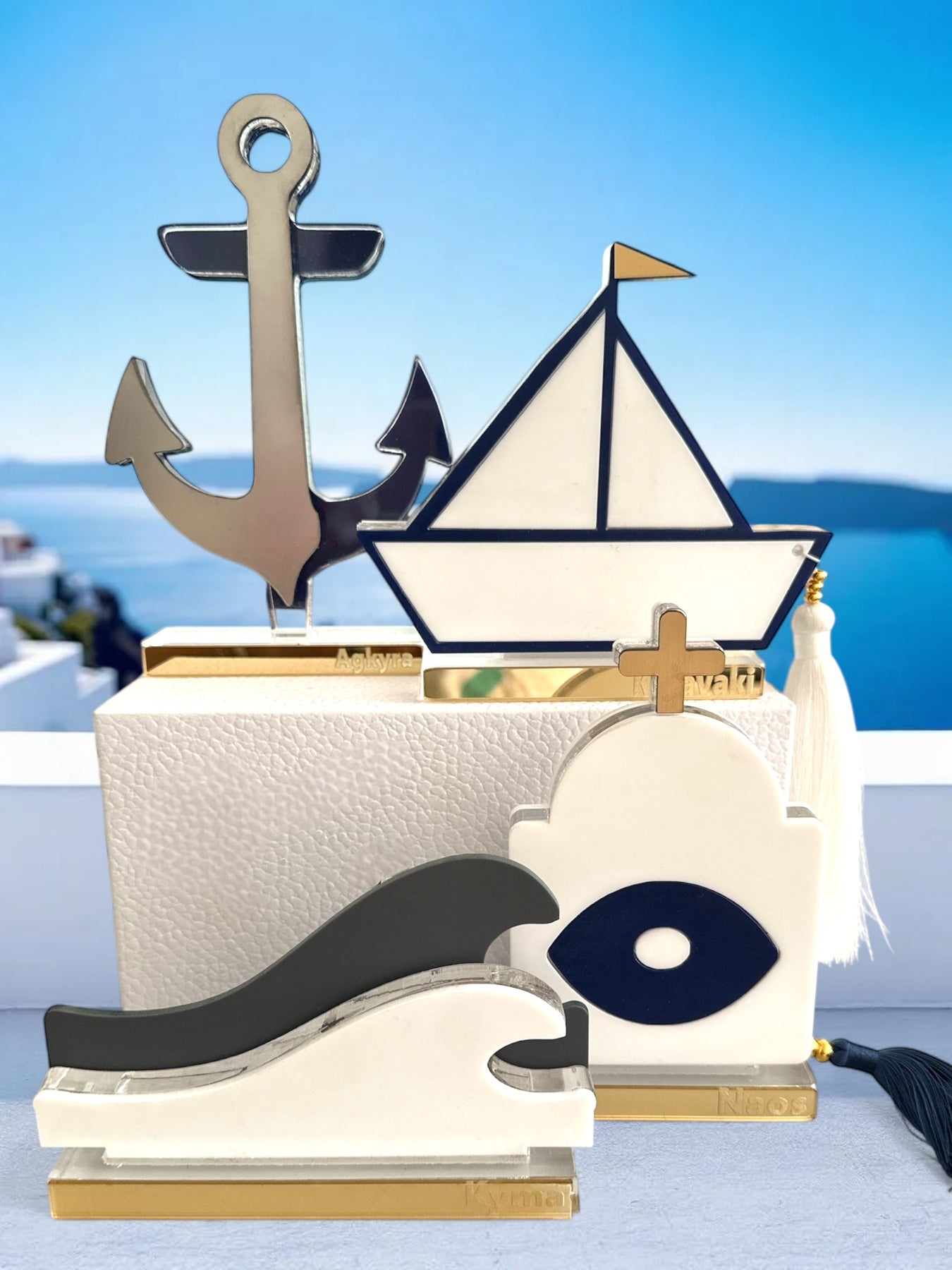 Σύγχρονα διακοσμητικά γούρια από πλεξιγκλάς σε κυκλαδίτικο φόντο: άγκυρα από χρυσό καθρέφτη, καραβάκι με navy περίγραμμα και φούντα, κύμα σε λευκό-γκρι, και ναός με ματ μάτι – όλα σε διάφανη και χρυσή βάση.