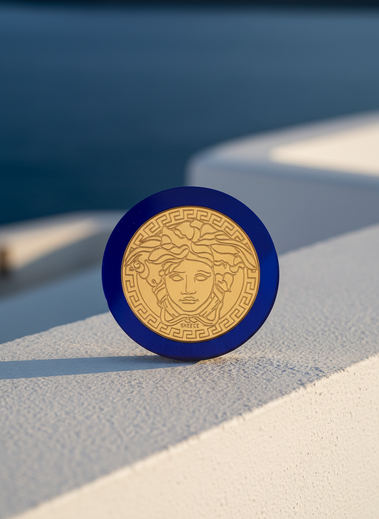 Στρογγυλό γούρι μέδουσα από μπλε πλεξιγκλάς καθρέφτη με χρυσή χάραξη, τοποθετημένο σε κυκλαδίτικο λευκό τοίχο με φόντο το βαθύ μπλε Αιγαίο.