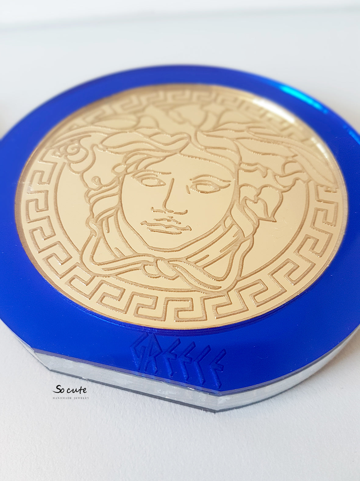 Κοντινή λήψη γουριού μέδουσα από μπλε πλεξιγκλάς καθρέφτη με χρυσή χάραξη και ελληνικό μοτίβο